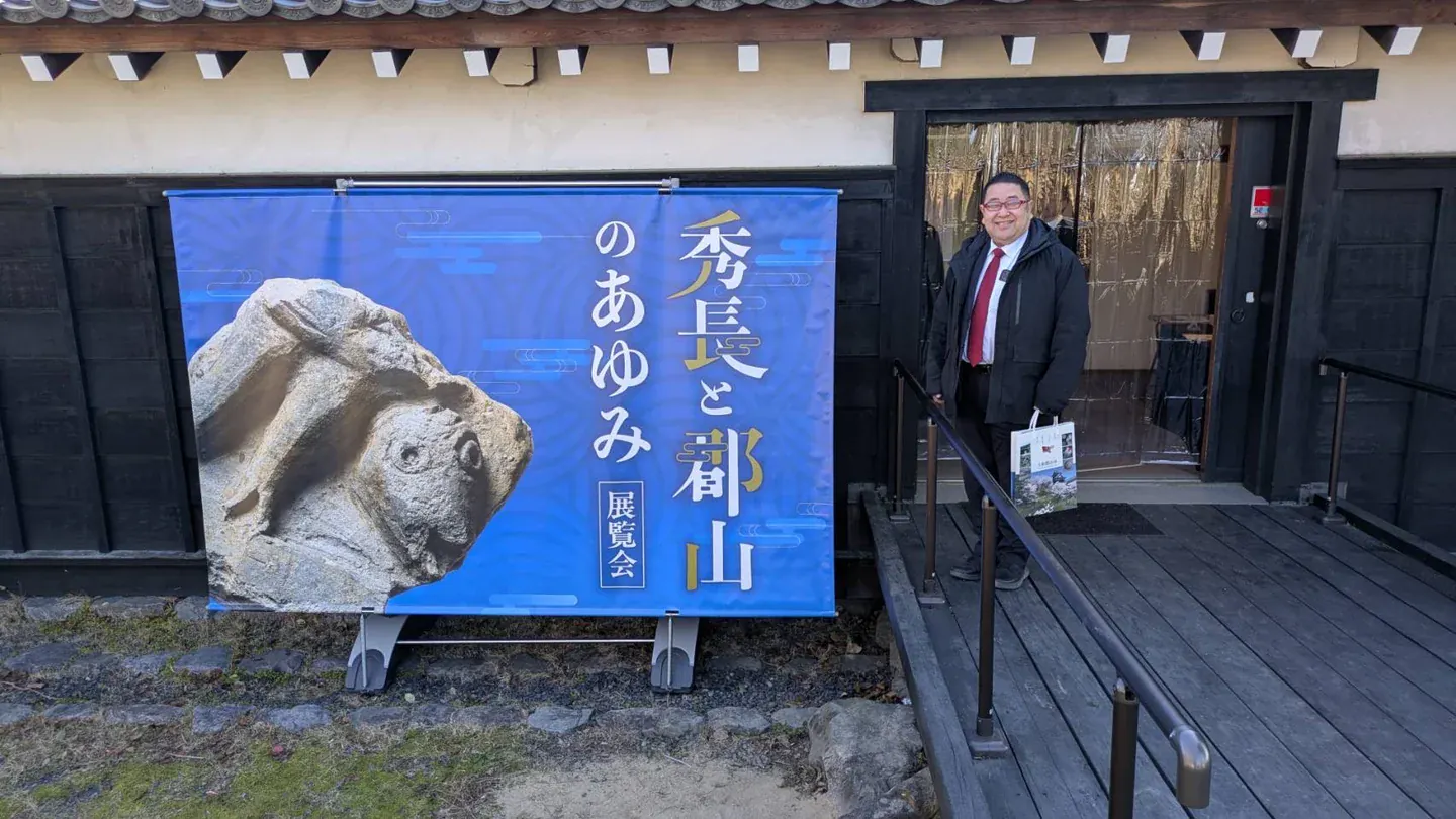 地域の展覧会の案内横に立つ鈴木たけし